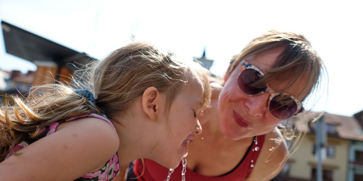 beautiful mid adult mother is drinking water with her daughter at outdoor water fountain in summer