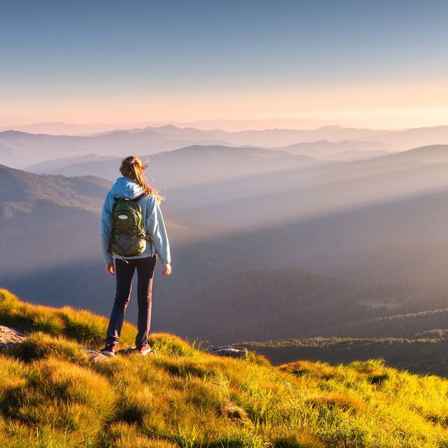 beautiful mountains in fog and standing young woman with backpack on the peak at sunset in summer landscape with sporty girl, green grass, forest, hills , blue sky with sunbeams travel and tourism