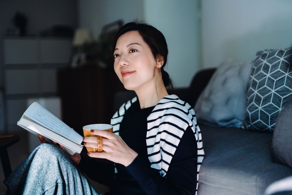 beautiful smiling young asian woman relaxing at home, reading a book with a cup of tea having a technology free moment enjoying a quiet time and relaxing moment at cozy home healthy lifestyle living
