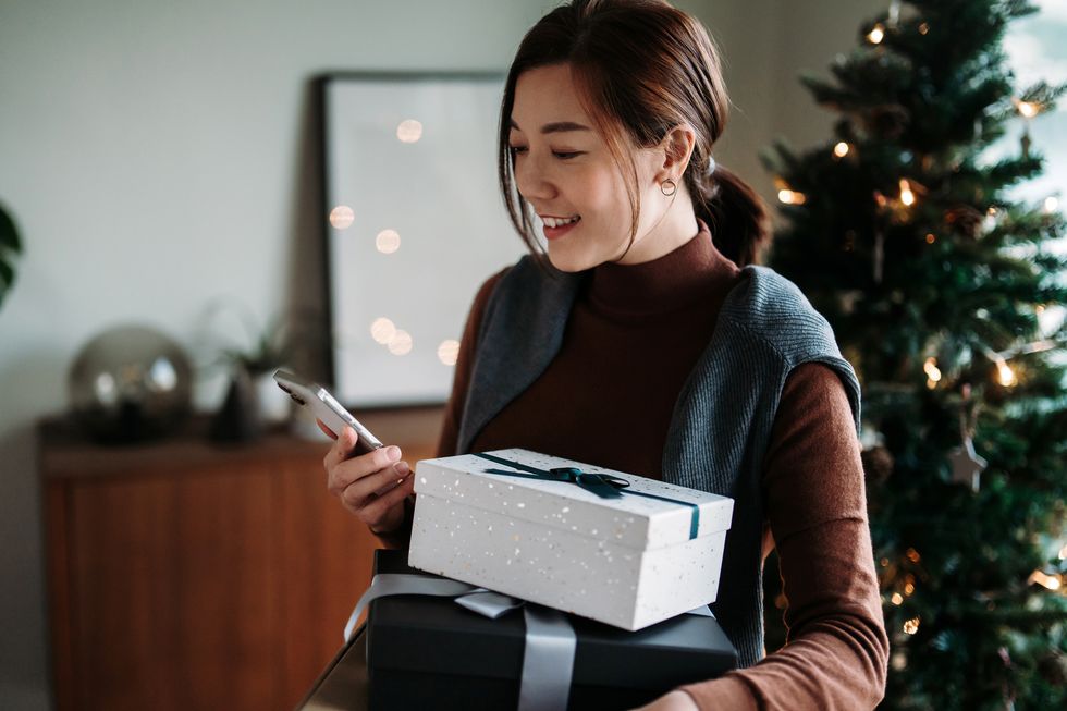 beautiful smiling young asian woman using smartphone while holding wrapped christmas presents, standing against decorated christmas tree christmas lifestyle christmas shopping receiving christmas gifts from friends and family holiday and festive vibes