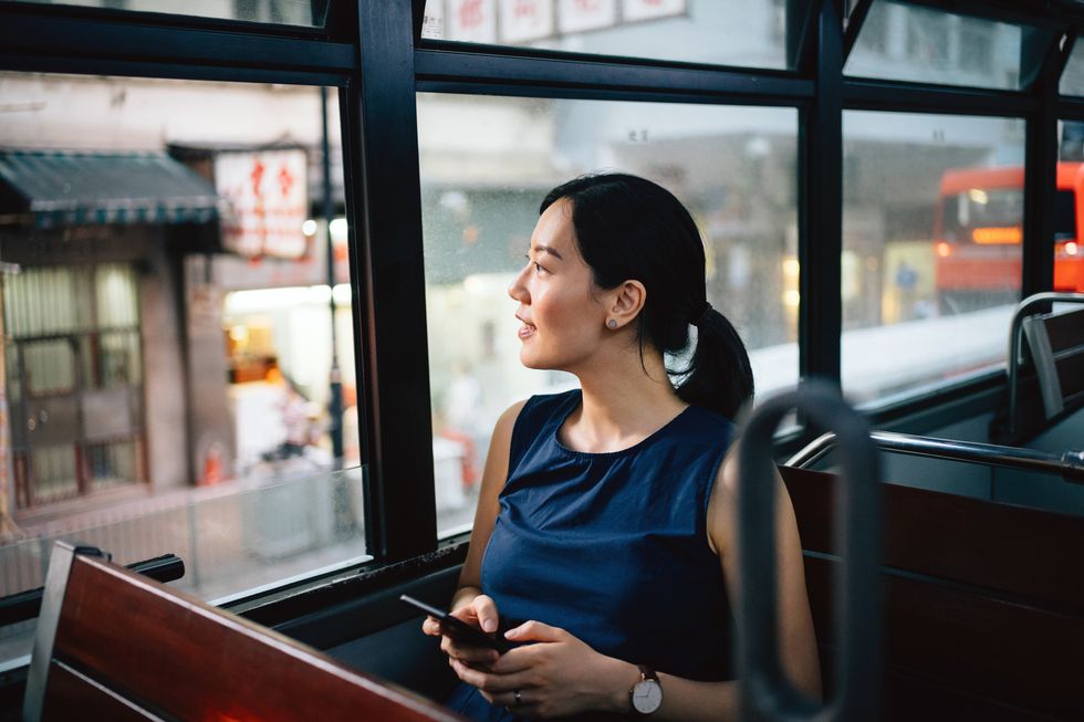 beautiful young asian lady with smartphone enjoying city scene through window while riding on public transportation in city