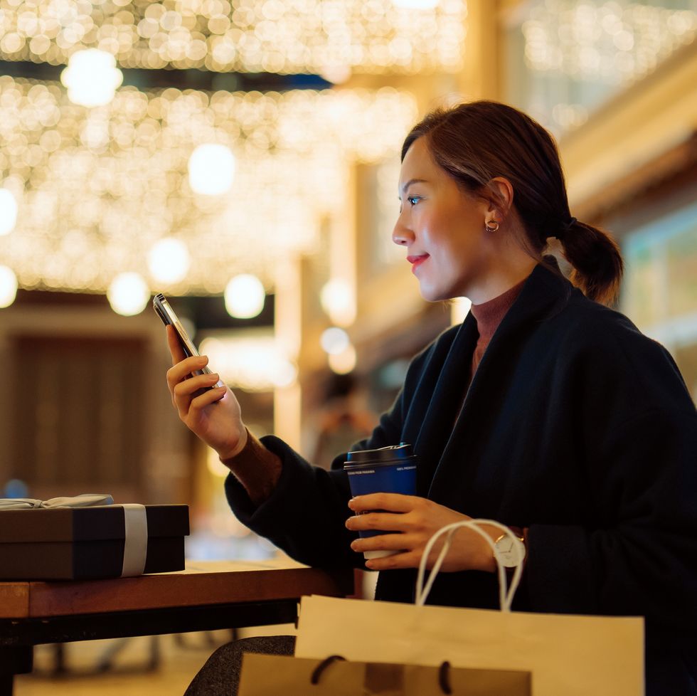 beautiful young asian woman relaxing on a bench and checking smartphone after shopping in the city, with decorated high street and illuminated christmas lights in background. enjoying christmas shopping. festive vibes. christmas is almost here