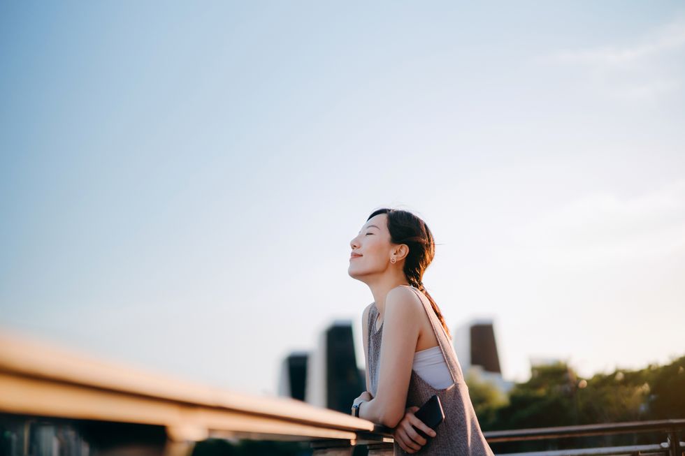 beautiful young asian woman with her eyes closed relaxing outdoors setting herself free and feeling relieved enjoying fresh air and the calmness with head up against sunlight at sunset