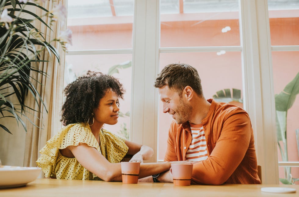 a beautiful young couple looks very much in love as they sit at a dining table sipping hot drinks and flirt