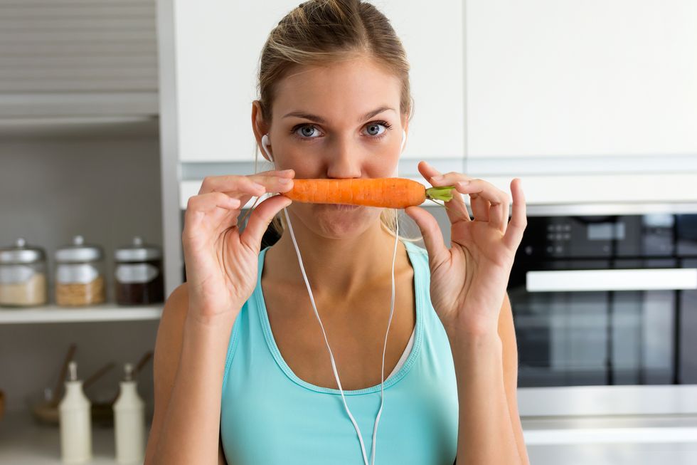 Beautiful young sporty woman playing with carrot while listening to music with earphones in the kitchen.
