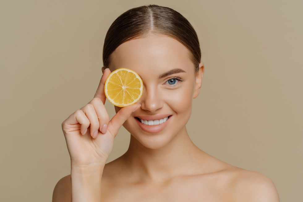 beauty and vitamin c concept headshot of lovely dark haired european woman smiles gently covers eye with slice of juicy lemon poses half naked against brown wall has healthy skin eats healthy food