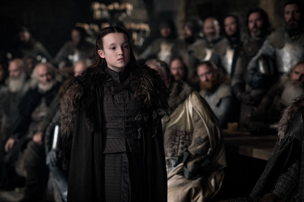 bella ramsey stands with her back to a large group of men, she is wearing a dark outfit with a fur accented long coat and has a stern look on her face