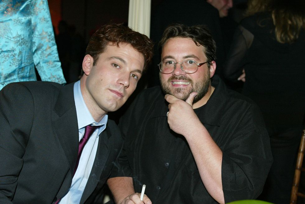 ben affleck, wearing a black suit and red tie, sitting next to a smiling kevin smith, who wears a black shit and glasses, holding a cigarette