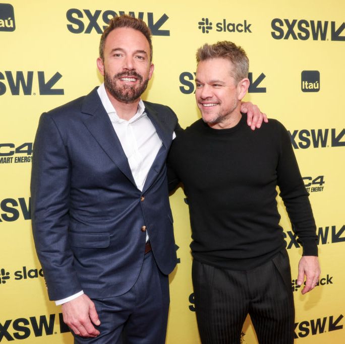 ben affleck, wearing a blue suit, smiles and puts his arm around the shoulder of a smiling matt damon, wearing a black shirt and pants, as they stand in front of a yellow wall with sxsw logos on it