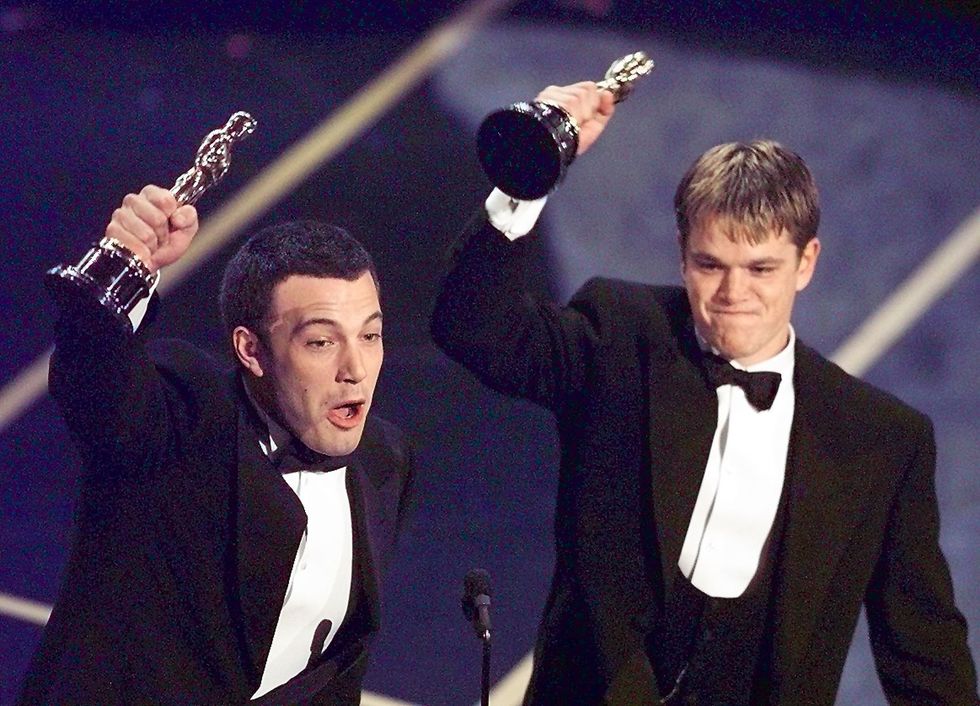 ben affleck and matt damon, both wearing black tuxedos, stand on stage in front of a microphone and hold academy award statuettes in the air with one hand each