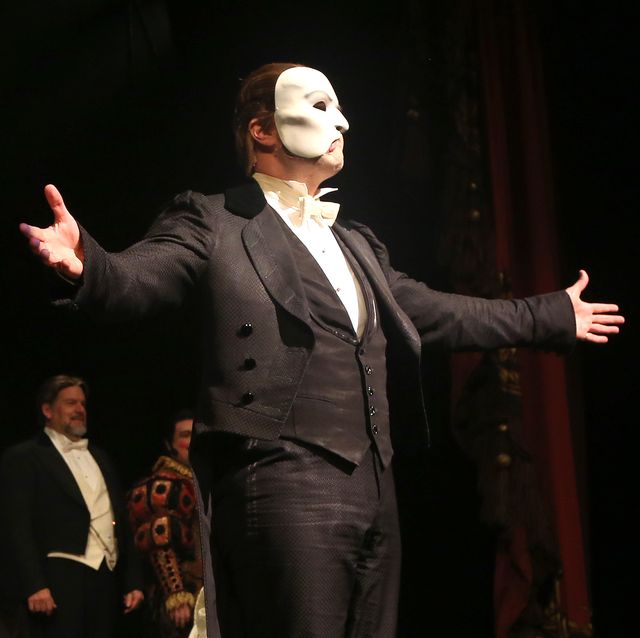 ben crawford dressed as the phantom of the opera, spreading his arms toward the audience while standing on a stage, with other actors standing behind him