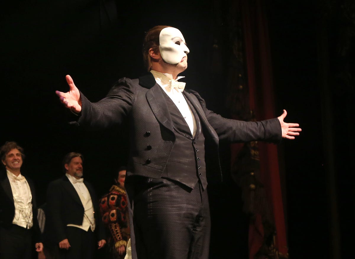 ben crawford dressed as the phantom of the opera, spreading his arms toward the audience while standing on a stage, with other actors standing behind him
