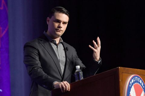 ben shapiro, wearing a black suit and gray shirt, standing at a podium on a stage and gesturing with his hand