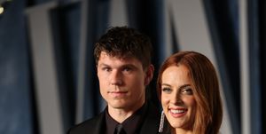 ben smith petersen and riley keough stand together and pose for a photo, he wears an all black suit and tie, she wears a glittering gold dress and smiles at the camera