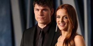 ben smith petersen and riley keough stand together and pose for a photo, he wears an all black suit and tie, she wears a glittering gold dress and smiles at the camera