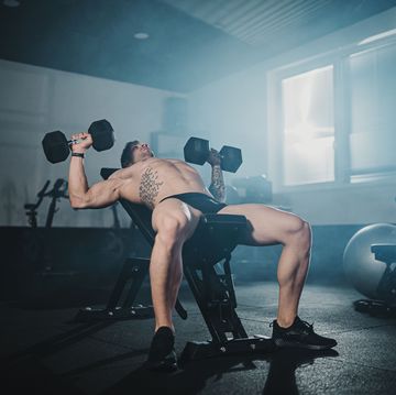 young male athlete doing strength training with dumbbells on bench at gym