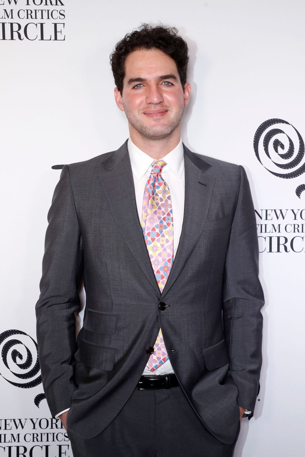 benny safdie smiles at the camera, he wears a gray suit with a white collared shirt and multicolored tie