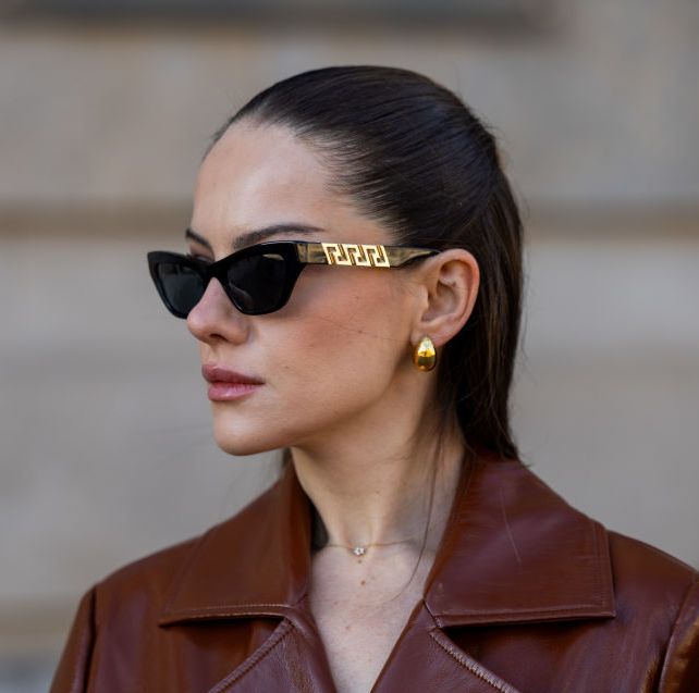 a brunette woman wearing black and gold sunglasses, gold earrings, and a burgundy leather trench coat on the street in berlin