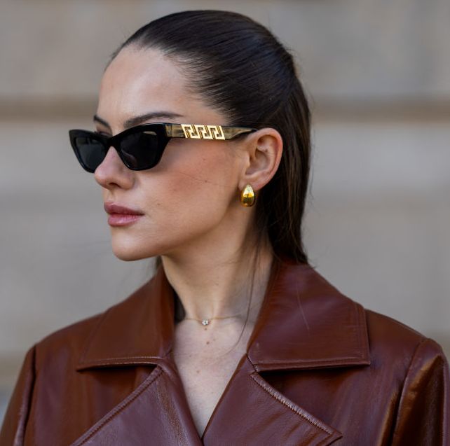 a brunette woman wearing black and gold sunglasses, gold earrings, and a burgundy leather trench coat on the street in berlin