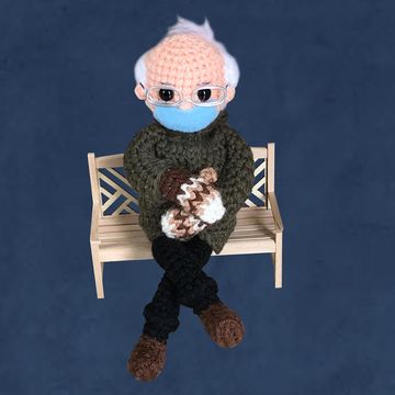 bernie sanders crochet doll sitting on a bench with brown mittens, face mask, and coat