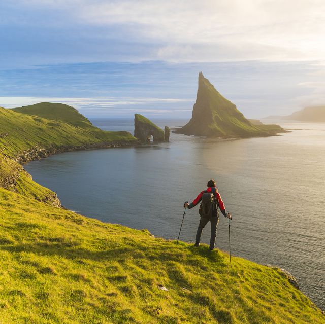 hiker on top of hill looks at the majestic drangarnir rock and the calm water of the ocean, vagar island, faroe islands, denmark