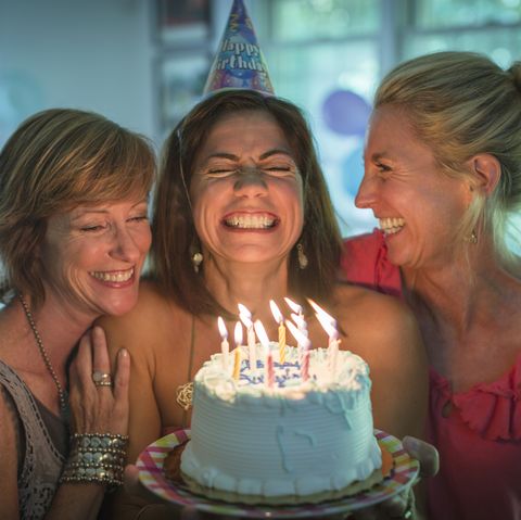 smiling woman holding birthday cake with lit candles making wish while two friends look on
