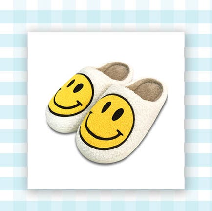 a pair of sherpa cream colored slip on slippers with smiley faces on them and a yellow and clear bag filled with lotion, body wash and body spray