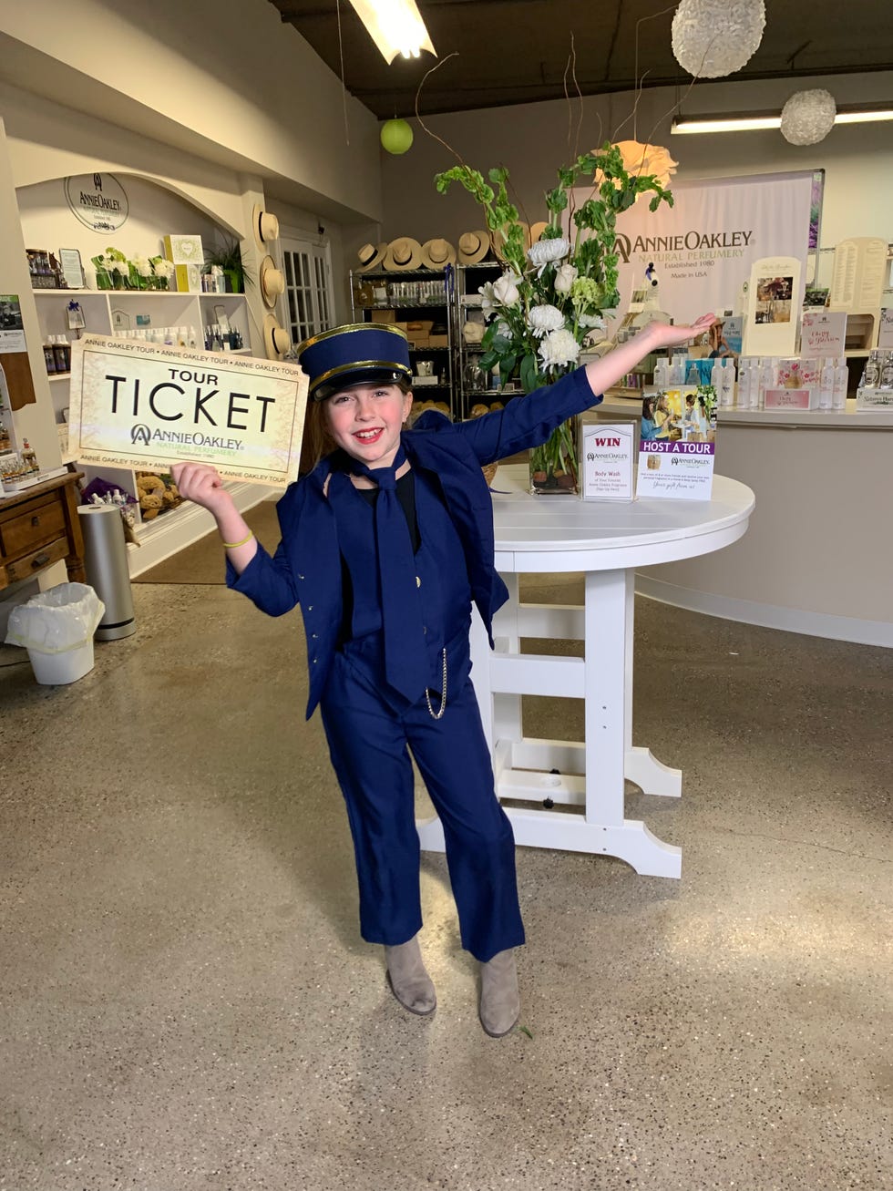 a person in a cobalt blue bellhop uniform with a ticket directing visitors to the annie oakley natural perfumery tour while standing in front of tables and shelves of perfume bottles the annie oakley natural perfumery tour is a good housekeeping pick for best factory tours