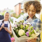 a young woman stands at the front door of her home and reads the card from a big bouquet of flowers  behind her is the delivery girl who has just dropped them off the shallow focus is on the bouquet blurring off to the people