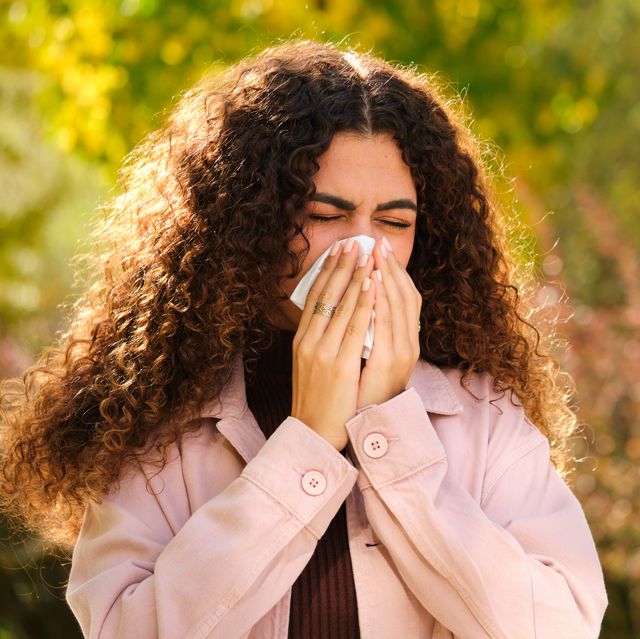 young woman with curly hair blowing your nose in a tissue on tree lined street