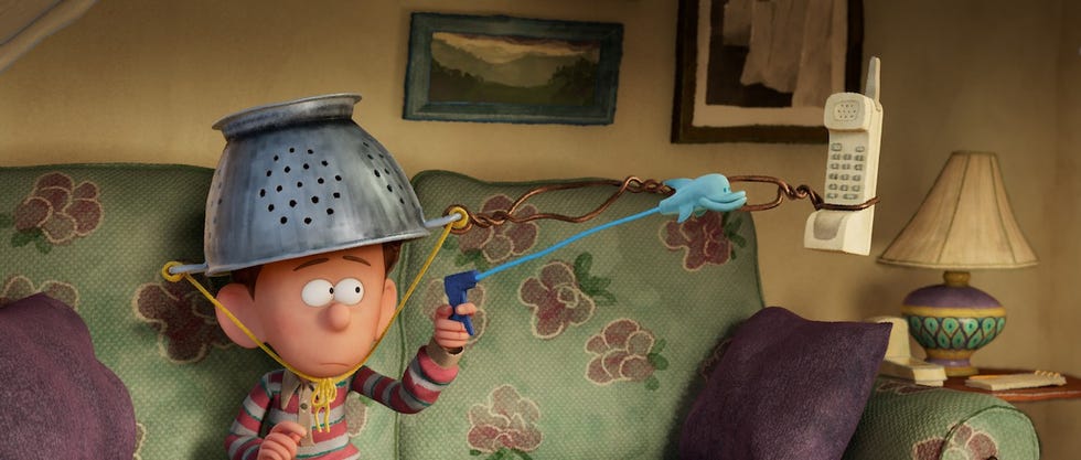 orion sits on a couch with a metal colander on his head and reaches for something in a scene from orion and the dark