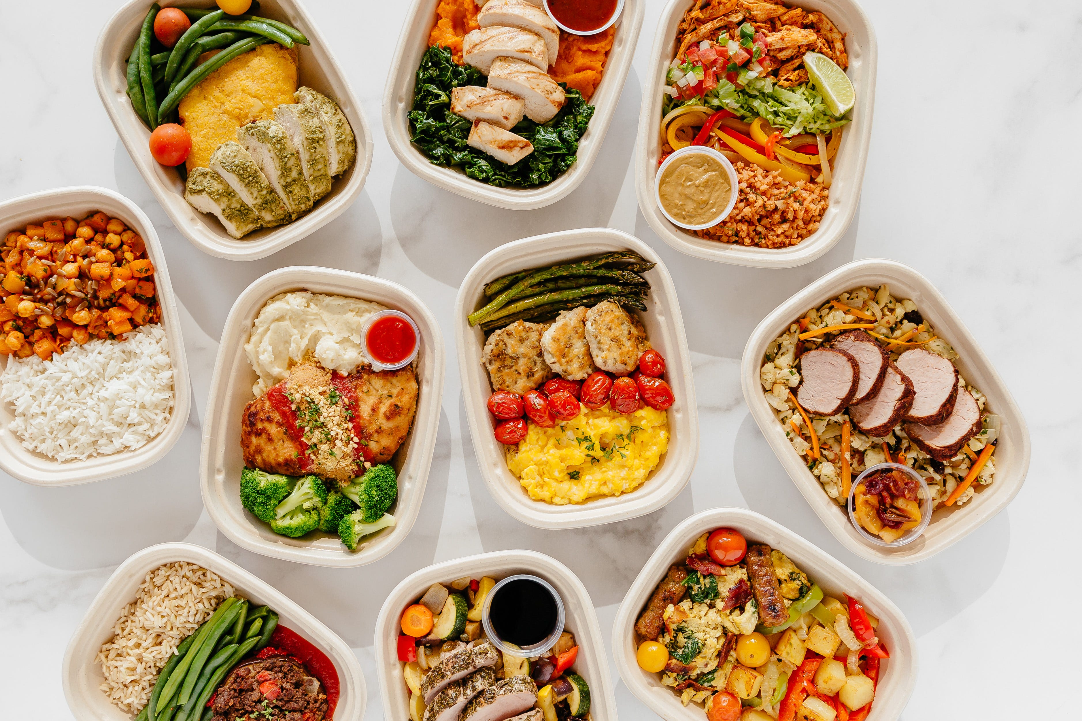 an assortment of meals including pork tenderloin and chicken entrees in meal prep containers on a white countertop from territory foods, a good housekeeping pick for best healthy meal delivery service