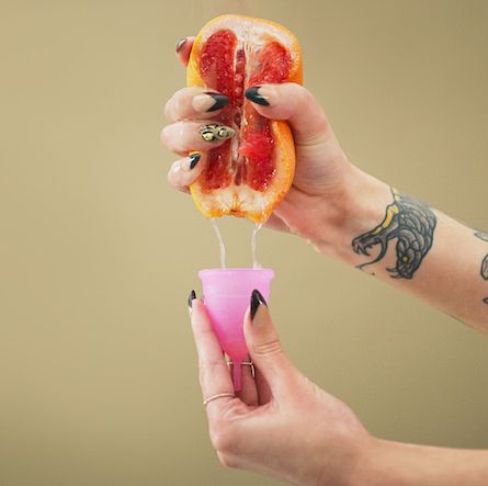 studio shot of a woman squeezing juice from a grapefruit into a menstrual cup against a brown background