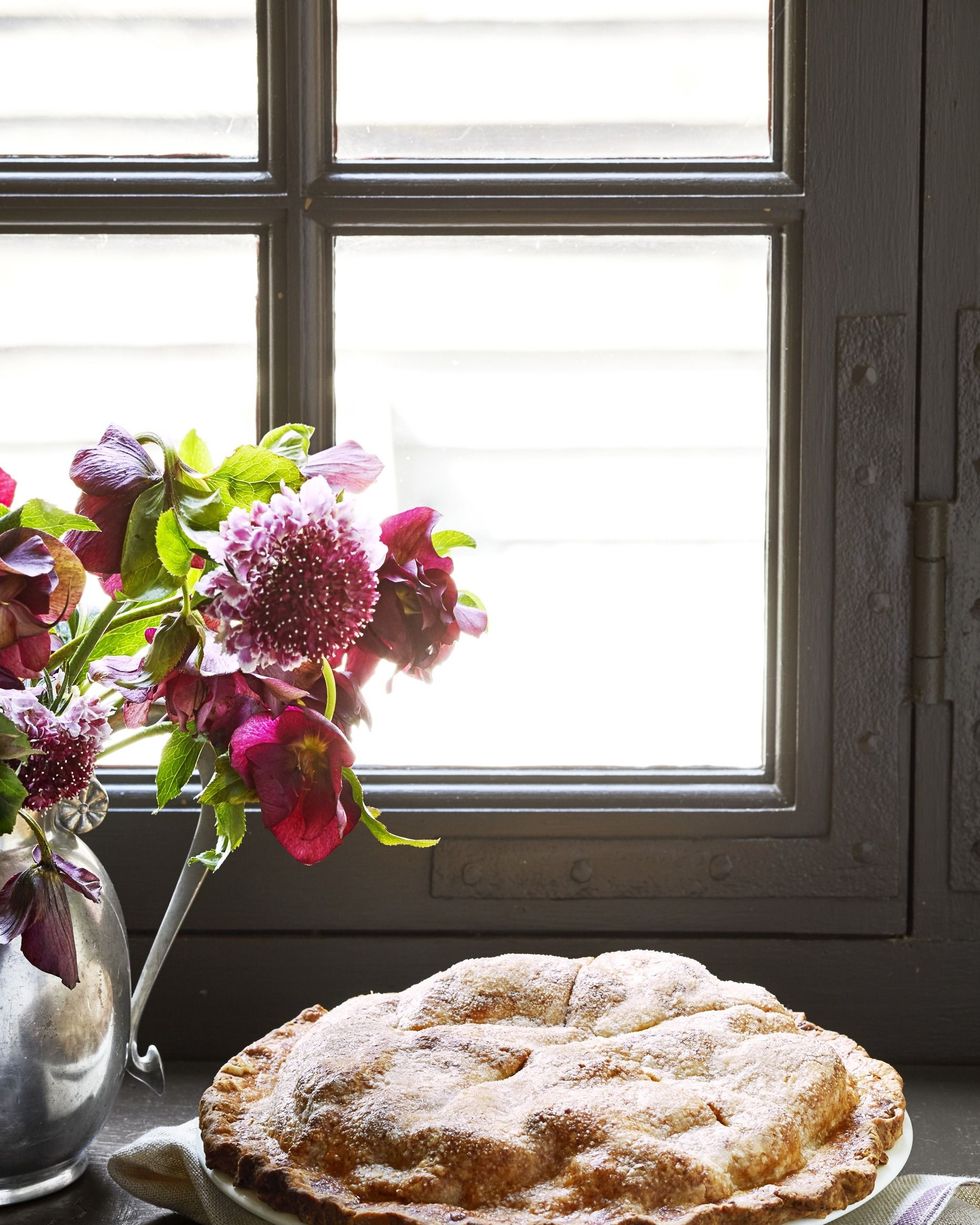 double crust apple cheddar pie in a white pie dish on a windowsill with a pie cutter and next to a metal vase of flowers
