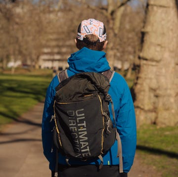 Best running backpack the back of a man stood in a park wearing a blue coat and cap, and a black running backpack with the words ultimate direction