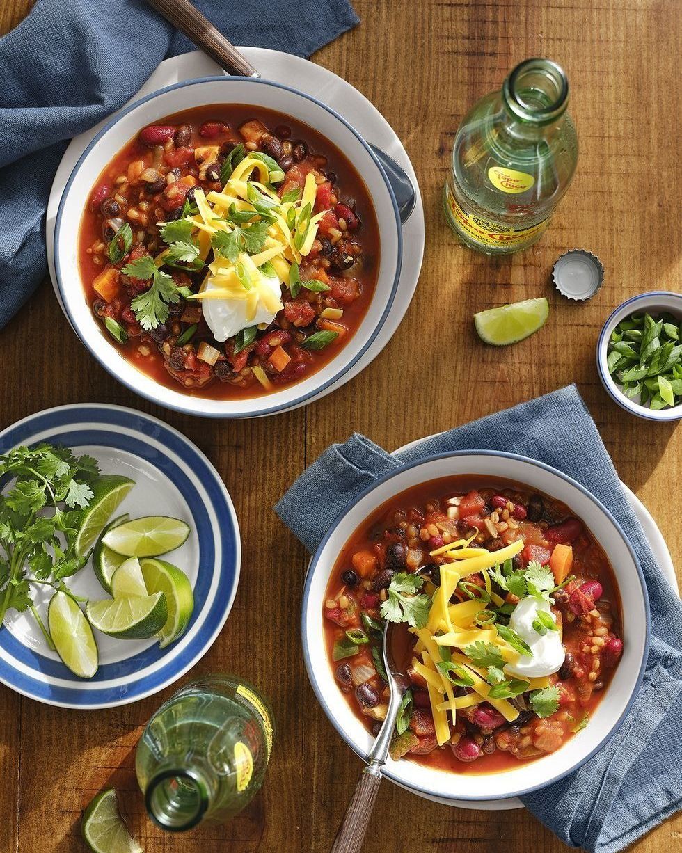 slow cooker vegetarian chili with grains and beans in two white bowls with blue trim topped with shredded cheddar and cilantro and sliced green onions and a dollop of sour cream