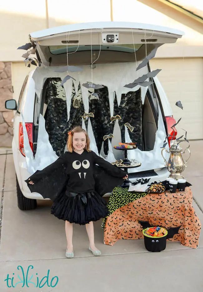 little girl in black bat costume with wings hanging between arms and waist and bat fangs and big silly eyes on shirt standing in front of trunk or treat with vehicle gate open to reveal cave backdrop with paper bats hanging in front of it and table with bat tablecloth with bat themed treats on it