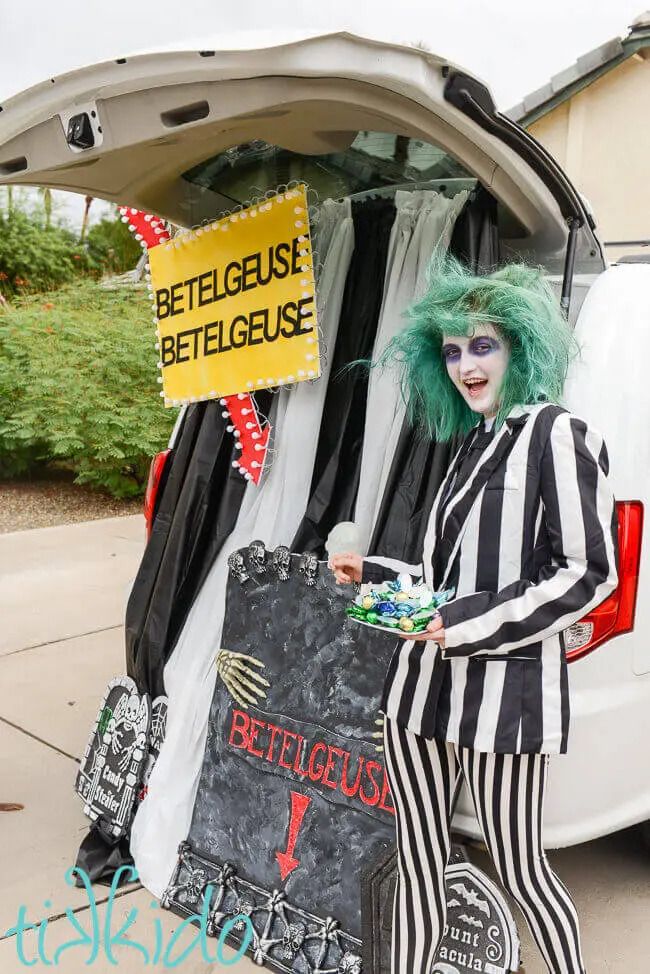 woman dress as beetlejuice in green wig and vertical black and white striped suit jacket and leggings holding plate of candy with black and white curtains in front of open tailgate of vehicle with betelgeuse sign with arrow and faux headstones