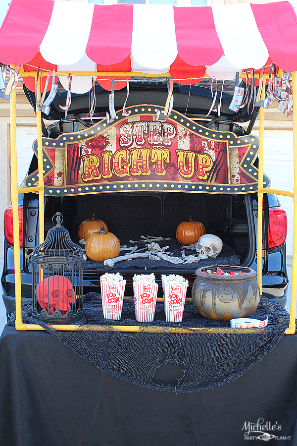 open tailgate with step right up carnival look banner and red and white striped canopy over top in front of it under the canopy are three red and white striped classic popcorn boxes with popcorn, a prop cauldron with candy, a red skull inside a black bird cage and inside the vehicle are pumpkins and prop bones