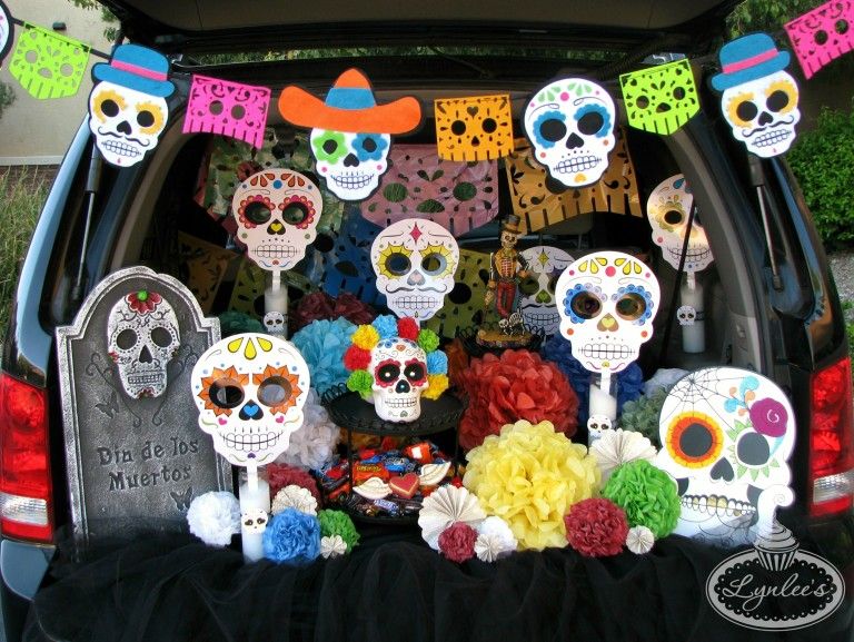 open tailgate of vehicle revealing colorful day of dead themed trunk or treat with sugar skull banner, stand up sugar skulls, paper flowers and a stand up gravestone with dia de los muertos written on it with sugar skull