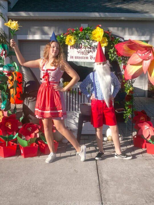 couple dressed as gnomes with man with a red triangular hat and long white beard and woman in red dress with short blue triangular cap in front of vehicle with giant faux red flowers around it, a white gate in front of it, which is beneath a garden arch with faux greenery and flowers
