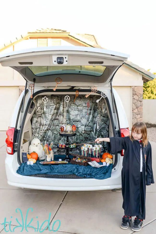 little girl dressed in harry potter black robe pointing a wand in front of a suv with the tailgate open revealing hogwarts themed items like hanging keys a white owl pumpkins, a curtain printed with a stone wall pattern as a backdrop and more