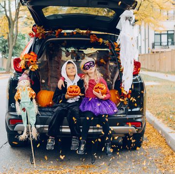 boy and girl holding jack o lanterns in open tailgate of parked vehicle with leaves blowing and on bunting at top of tailgate there is a fabric scarecrow on a stick leaning against vehicle the girl is wearing a black mask and purple tutu