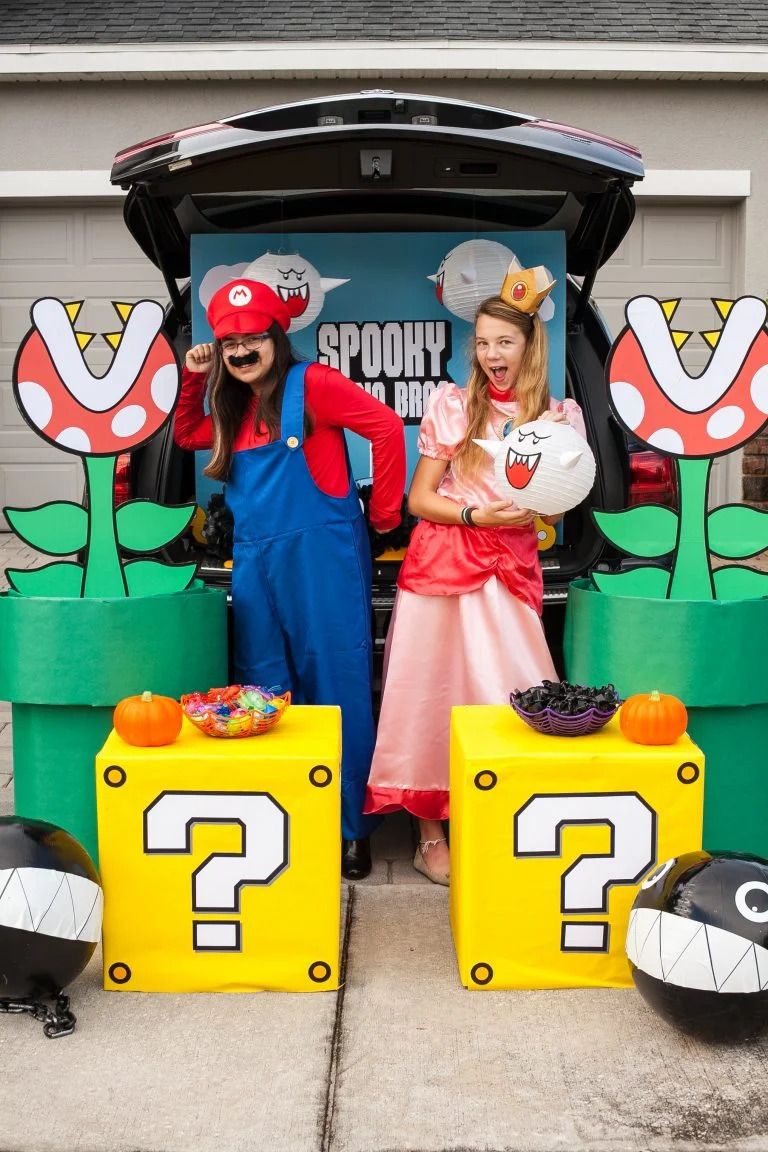 girl dressed as mario in sper mario brothers with mustache blue overalls and red hat and other girl dressed as princess peach in a pink and red dress with a crown they are standing behind props of two yellow boxes with question mark symbols on them, two black balls with teeth and eyes and cartoonish green piranha plants with red and white dotted heads and big mouths with teeth