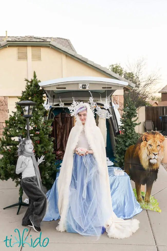 narnia trunk or treat with fur coats hanging in back of van, cut outs of snowflakes hanging from tailgate, cut out of a lion beside the van, on the other side is a faux christmas tree and a lamppost and there is a teen girl dressed as the white witch in a white fur trimmed cape and blue tulle skirt and a little girl dressed as the fawn in fur pants and a long scarf