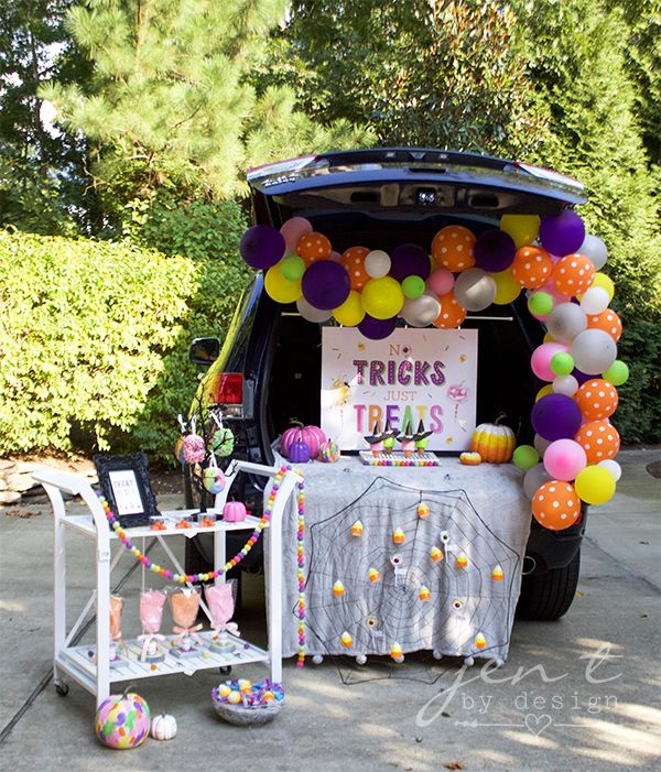 open tailgate decorated with colorful balloons, a sign reading no tricks just treats multi colored pumpkins and a white cloth hanging off the end covered in a faux spiderweb and candy corn lights there is a white cart next to the vehicle with cotton candy on stands a treat yo'self sign and a donut tree