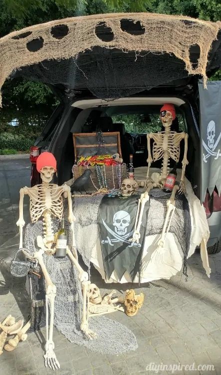 open tailgate of vehicle with a skeleton it, a skull, a treasure chest, a skull, prop bottles of rum and a pirate flag with skull and sword crossbones hanging over the back in front of the vehicle is another sitting skeleton, bones and skulls