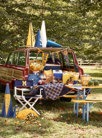 woody station wagon with tailgate open with blue and gold pennants hanging from the top, a football, a little cooler, yellow flowers a blue and yellow checked tablecloth and more are on the tailgate and there is a michigan director's chair and a picnic table that's blue and yellow in front of it