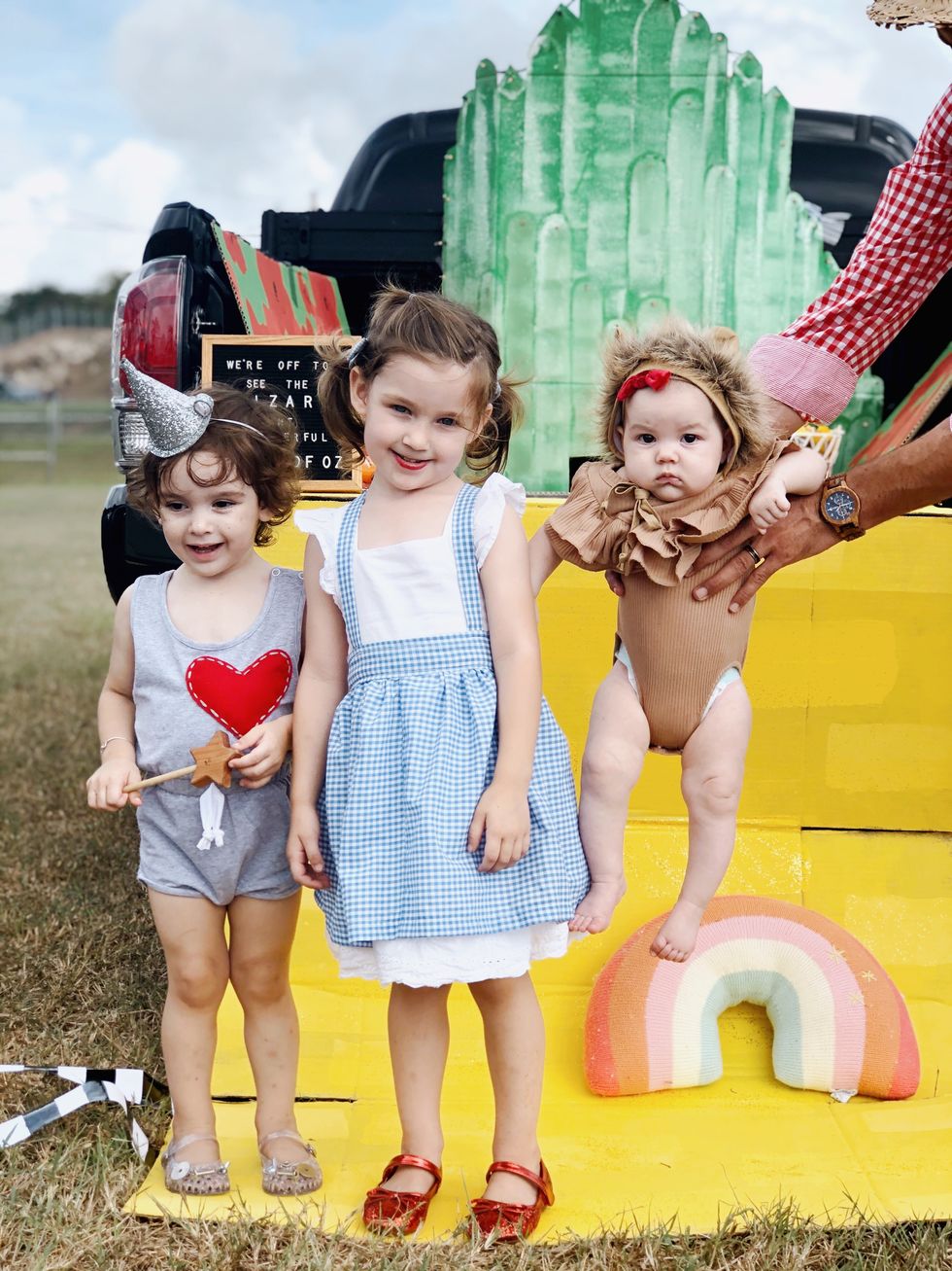 little girls dressed as dorothy in blue and white checkered pinafore dress and red shoes next to younger girl with wand and silver witch hat and baby dressed in lion onesey being held into the frame in front of truck with yellow painted brick road, rainbow pillow and painted emerald city in back and spilling out of tailgate
