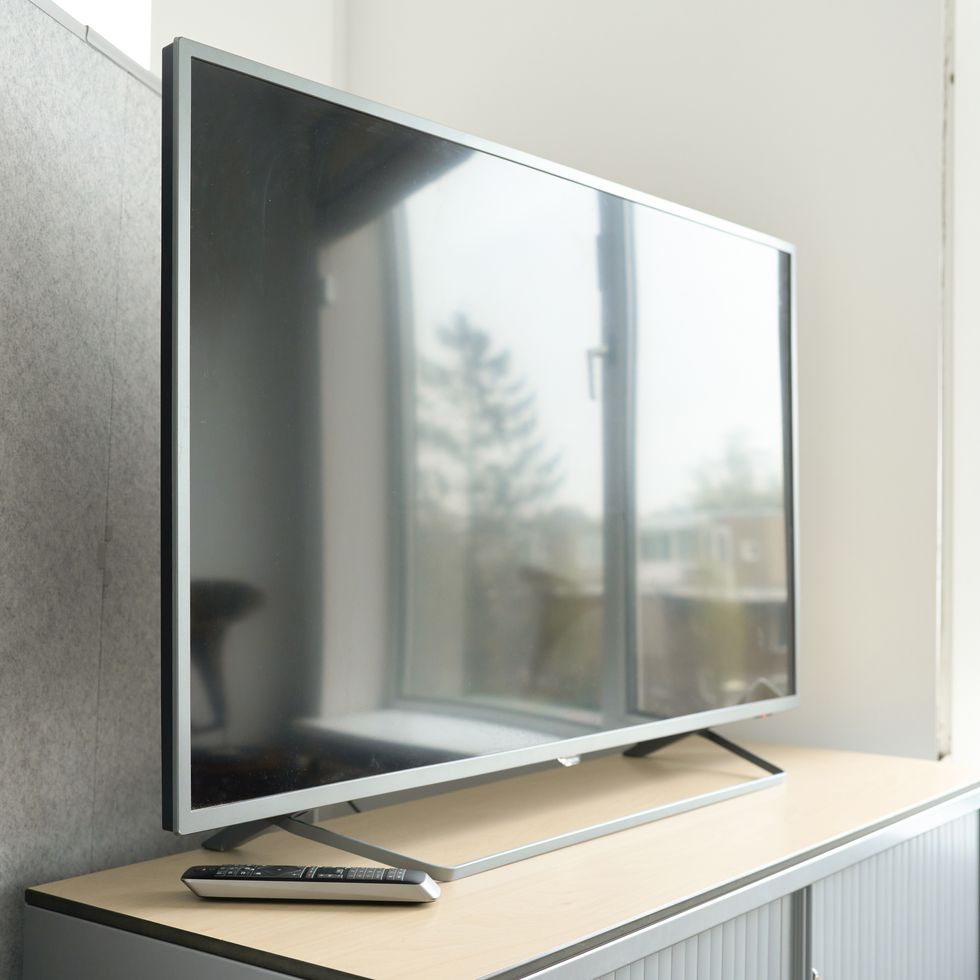 a large flat screen tv is turned off, but an image of a window is reflected, it is on a table where there is a control, in the background is a gray wall